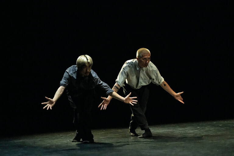 Twee mannelijke dansers staan met hun benen gekruisd en armen wijd op een donker podium.