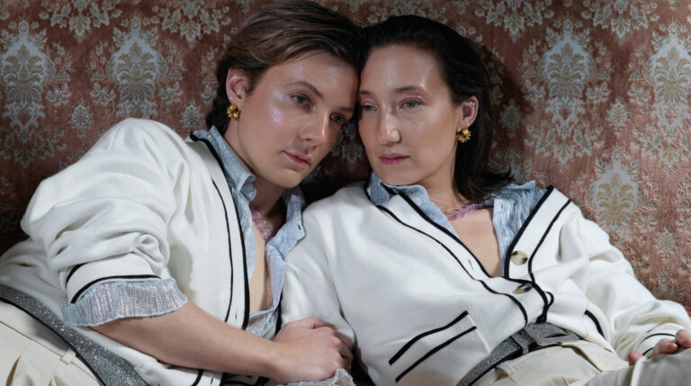 Twee acteurs die veel op elkaar lijken in dezelfde voornamelijk witte outfits liggen onderuit gezakt op een antieke bank.