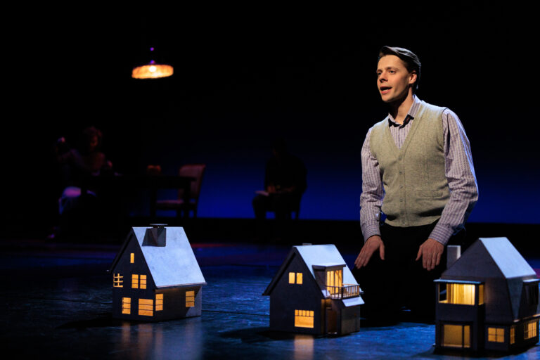 Een man staat op een donker toneel naast verlichte miniatuurhuizen.