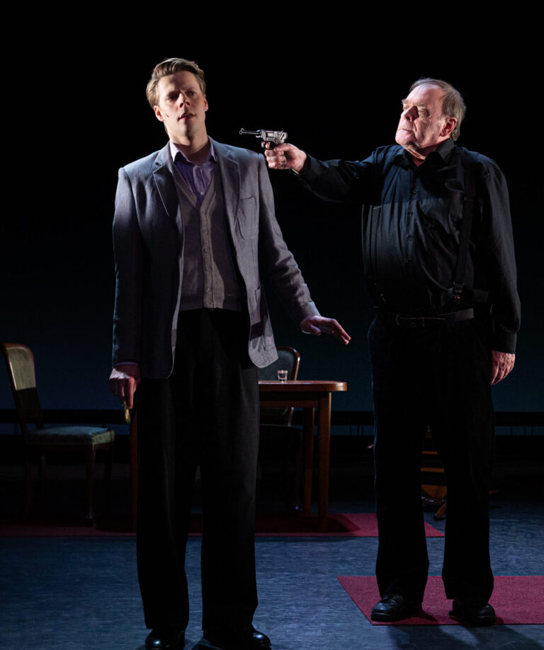 Op een donker podium staan twee mannen; de linker man draagt een licht pak en de rechter man in donker wijst naar hem.