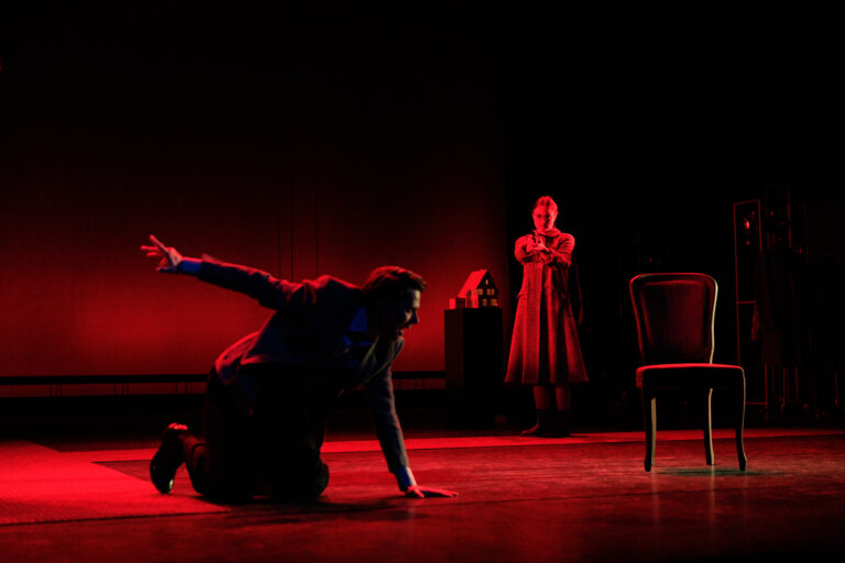 Op een rood verlichte theatervloer staan twee personen: één ligt op handen en knieën en de andere staat in een lange jurk, met een stoel op het podium.