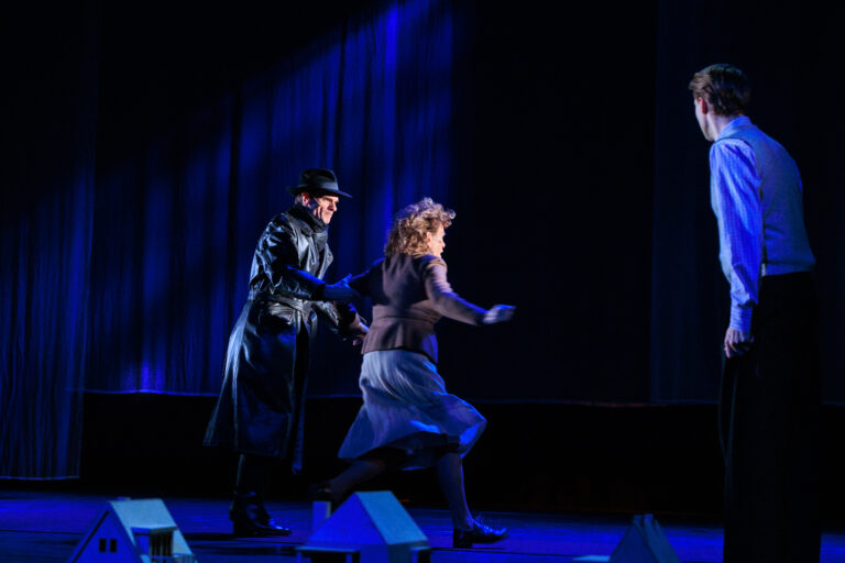 Twee dansers bewegen zich over een donker podium onder blauwe verlichting.