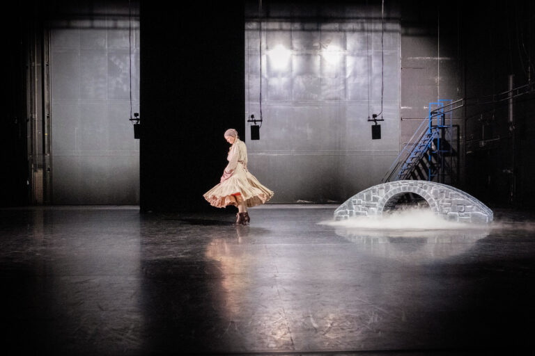 Vrouw staat in haar eentje op een minimalistisch podium, naast haar is een kleine brug decor