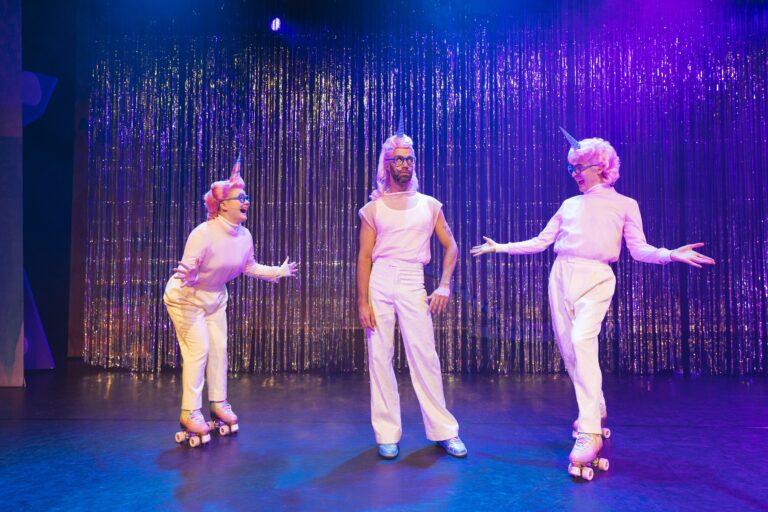 Drie performers in glimmende outfits en op rolschaatsen staan op een podium met een glinsterende achtergrond.