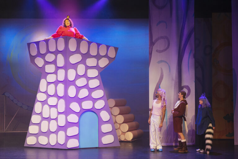 Een theatrale scène met een grote toren in felgekleurde uitvoering, waar een persoon bovenop zit, terwijl drie andere acteurs naar de toren kijken.