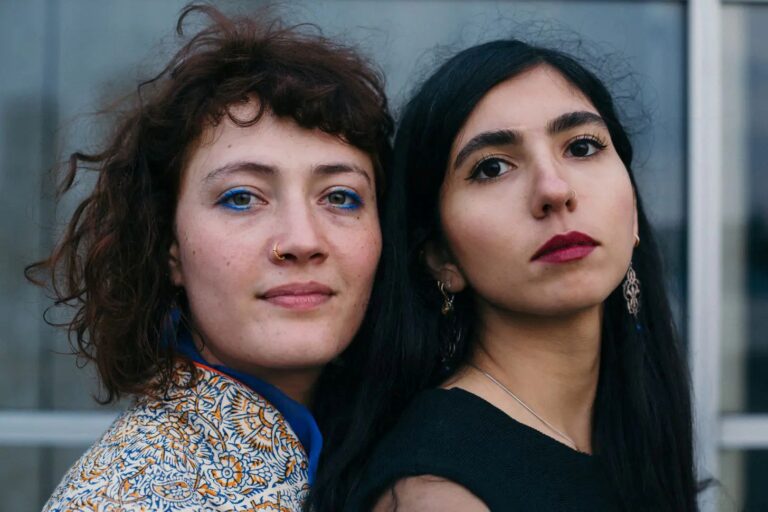 Twee vrouwen staan met hun ruggen tegen elkaar in een stedelijke omgeving, met verschillende kapsels en make-up.