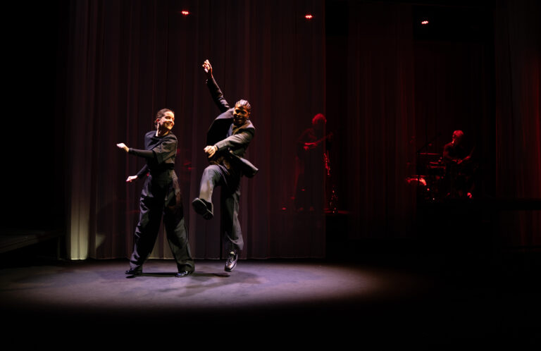 Twee dansers voeren een dansuitvoering uit op een donker podium, met een rood gordijn op de achtergrond.