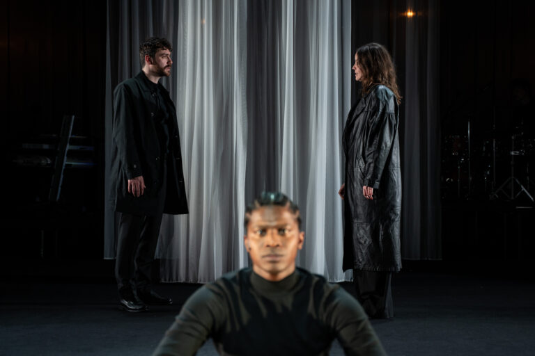 Drie personen op een donker podium met verticale gordijnen; de man in het midden staat dichter bij de camera terwijl de twee anderen op de achtergrond staan.
