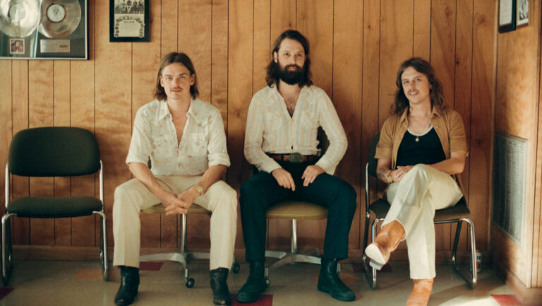 Drie muzikanten in retro kleding zitten in een wachtkamer met houten lambrisering en platen aan de muur.