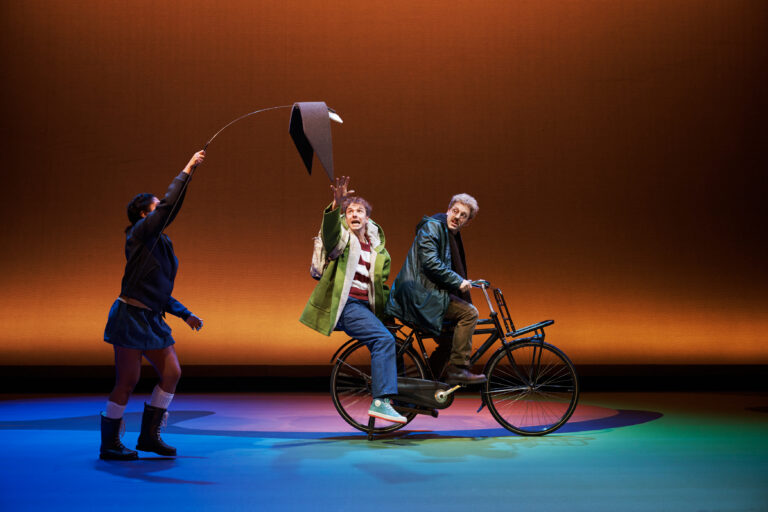 Drie personages op een kleurrijk podium, met een vrouw die een hoed boven een fietser uitsteekt terwijl een andere vrouw kijkt.