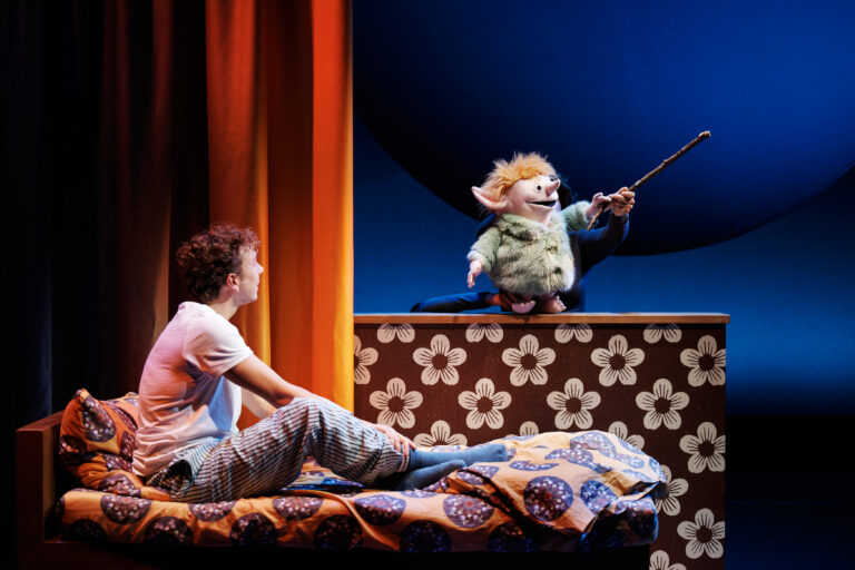 Een jonge man zit op een bed terwijl een pop met een toverstok hem toespreekt op een kleurrijk podium.