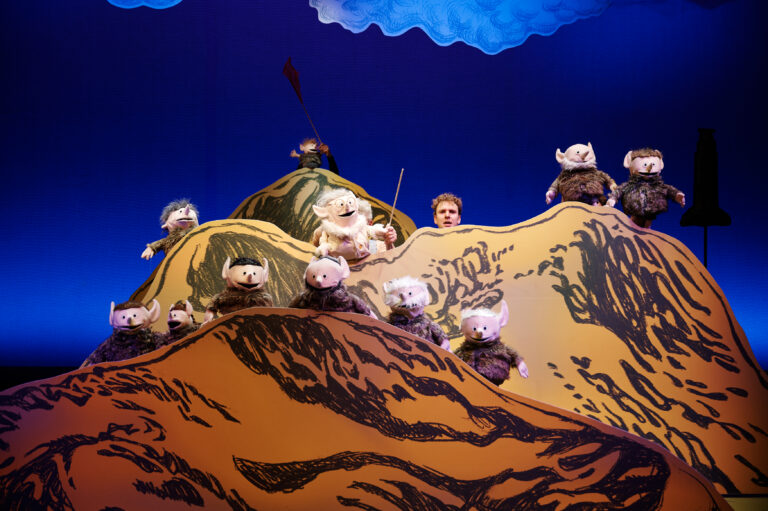 Een kleurrijk podium met heuvels en een groep poppen die de scène bevolken, terwijl een personage in het midden staat.