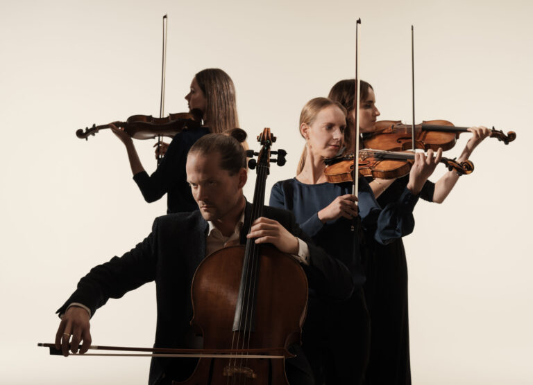 Het Dudok Quartet Amsterdam. Op de voorgrond zittend en iets van opzij gefotografeerd de cellist achter zijn cello, de strijkstok in de aanslag. Achter hem stand in verschillende posities de drie violistes, de strijkstokken ophoog wijzend. Alle vier de musici hebben de blik naar de linker of rechter buitenkant