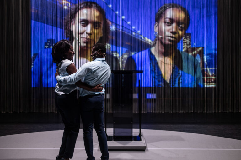 Twee acteurs, die Barack en Michelle Obama voorstellen, omarmen elkaar op het podium, terwijl op de achtergrond een grote projectie van de gezichten van hun dochters, Sasha en Malia Obama, te zien is.