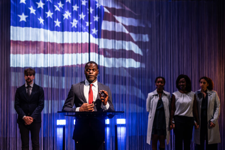 Een acteur, verkleed als Barack Obama, spreekt vanaf een podium met een projectie van de Amerikaanse vlag op de achtergrond. Achter hem staan drie andere acteurs, die Michelle Obama en hun twee dochters voorstellen, terwijl ze naar de toespraak luisteren.