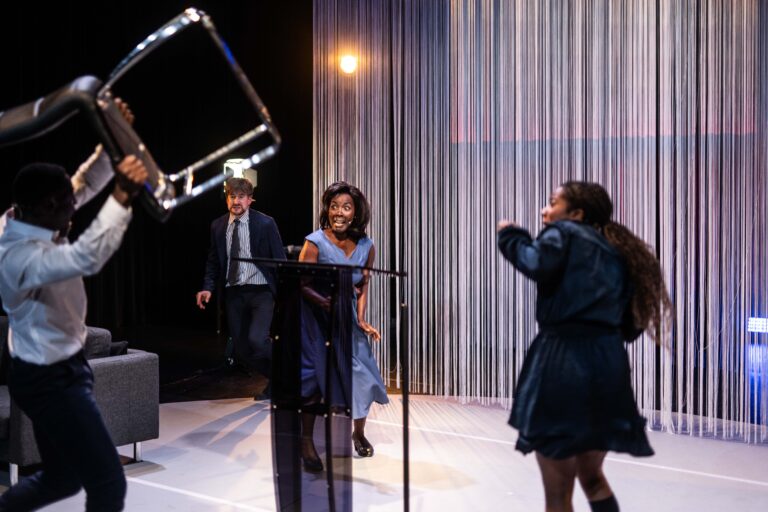 Een intense scène waarin een acteur, verkleed als Barack Obama, met een stoel in de lucht staat. De acteur die Michelle Obama speelt, kijkt angstig naar de situatie. Achter hen staan andere personages die zichtbaar betrokken zijn bij de hectische gebeurtenis.