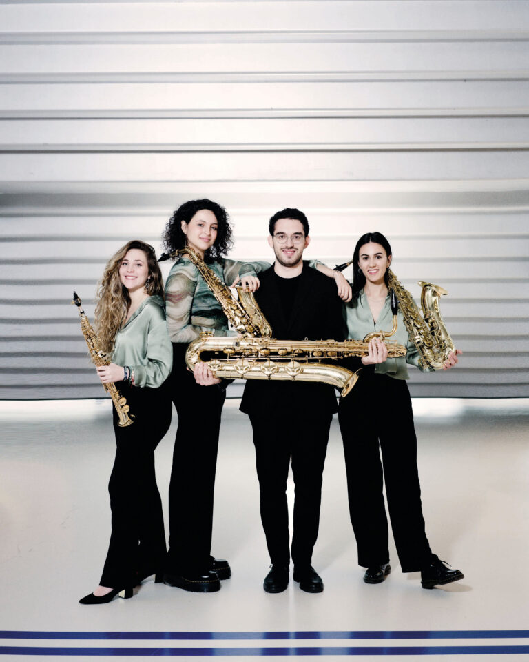 Drie jonge vrouwen met saxofoon en een man in zwart pak met lichtgrijze horizontale lijnen in de achtergrond