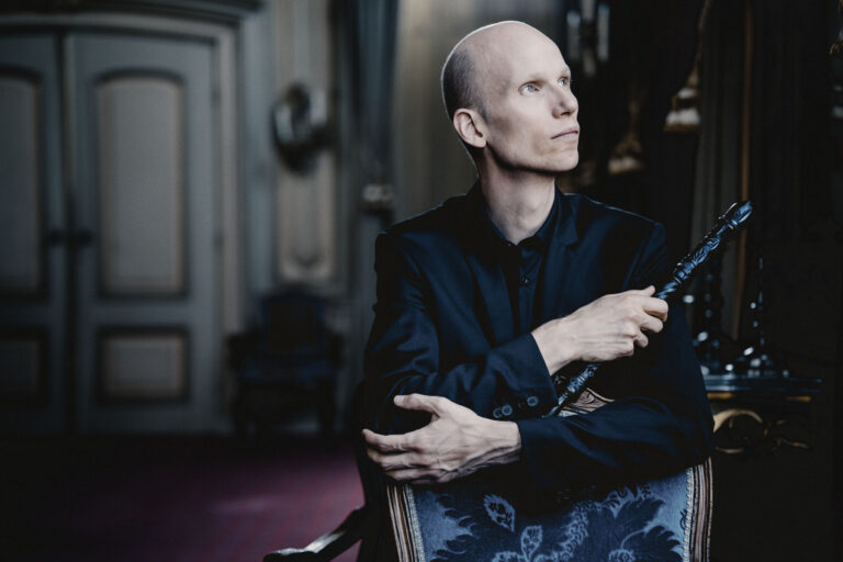 Een balding man in een zwarte outfit zit op een luxe stoel en houdt een fluit vast, terwijl hij naar de zijkant kijkt.