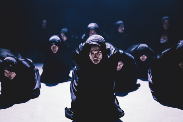 Dansers in volledige zwarte doeken gewikkeld en schmink op hun gezicht zitten gehurkt op het podium