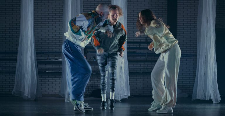 Drie dansers in kleurrijke, sportieve kleding staan in beweging op een podium, met blauwe gordijnen op de achtergrond die de ruimte een intieme sfeer geven.