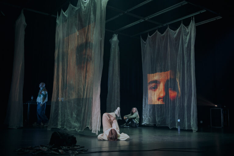 Op het podium hangen doorschijnende doeken waar gezichten op worden geprojecteerd, terwijl twee performers in ontspannen poses op de grond zitten, wat een intieme en dromerige sfeer oproept.