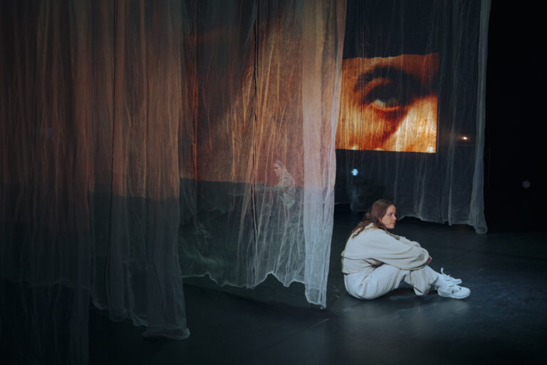 Een performer zit op de grond, omringd door doorschijnende doeken met projecties van ogen, wat een mysterieuze en contemplatieve sfeer oproept.