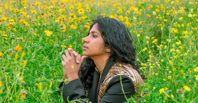 Een vrouw met lang, donker haar knielt in een veld met gele bloemen en kijkt dromerig naar de verte.