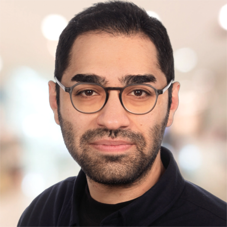 Een man met een baard en een bril kijkt recht in de camera, met een ingetogen achtergrond.