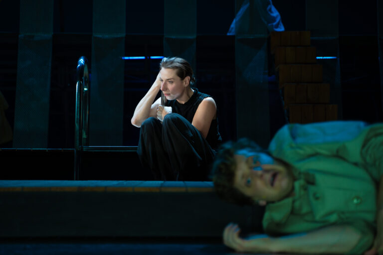 Vrouw leunt tegen een zwembad trapje aan met haar hoofd en kijkt beteuterd, op de voorgrond ligt een man op de grond met een angstige blik