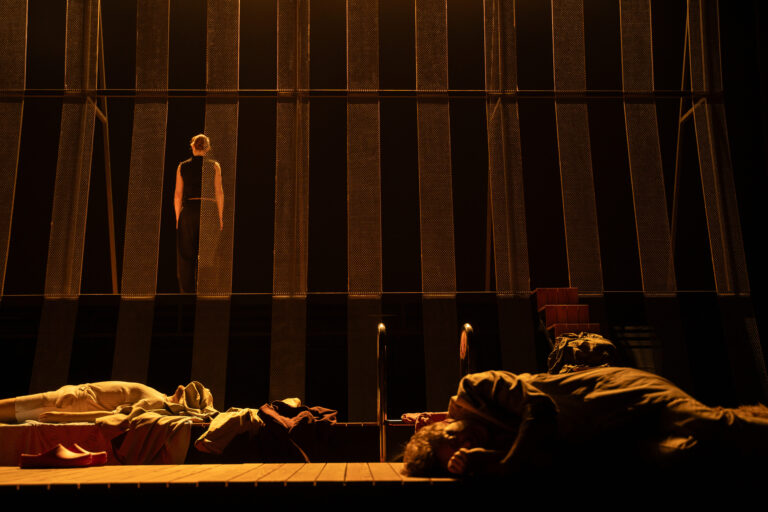 Er liggen verscheidene lichamen en kledingstukken op het podium, een vrouw staat belicht met haar rug toegekeerd op de achtergrond