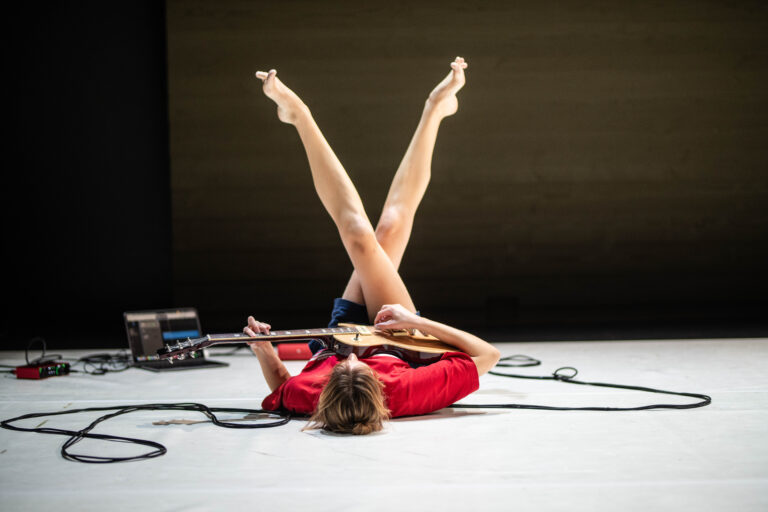 Annabel Koele ligt op de grond met de benen in de lucht en speelt een elektrische gitaar tijdens een voorstelling.