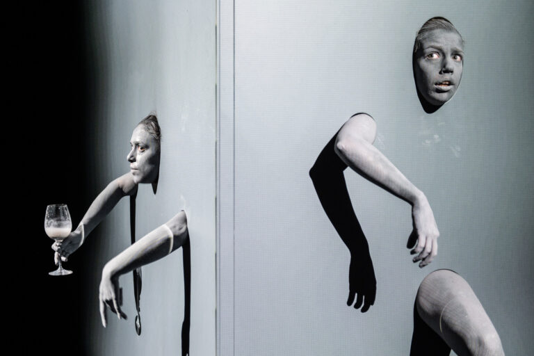 Twee performers, grijs geschilderd en deels door een muur gestoken, met een van hen die een wijnglas vasthoudt en de ander met een verbaasde uitdrukking