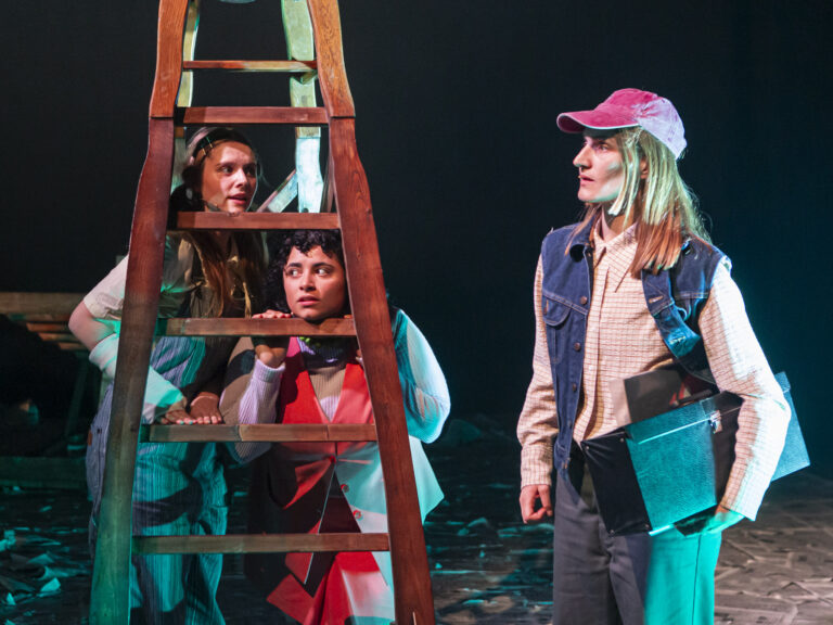 Drie acteurs staan rond een houten ladder in een theatrale setting, met een donkere achtergrond en sfeerverlichting.