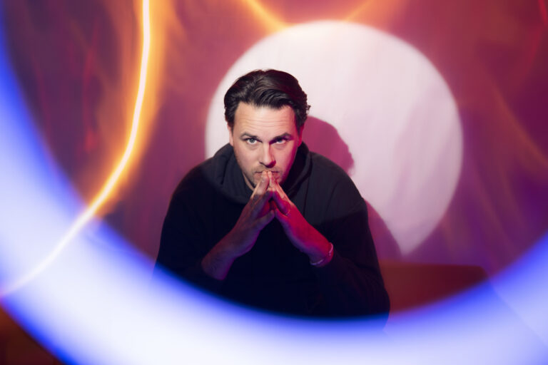 Man zit met handen tegen elkaar voor zijn mond en heeft verschillende lichten in roze, geel en blauw om hem heen.