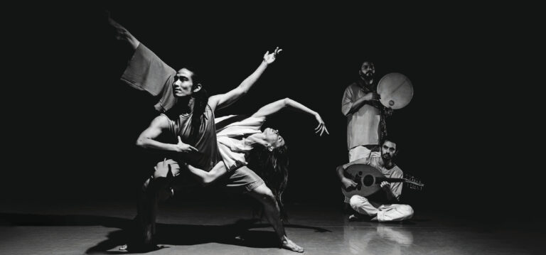 Zwart-wit foto van twee dansers die om elkaar heen dansen met op de achtergrond twee muzikanten met traditionele instrumenten.