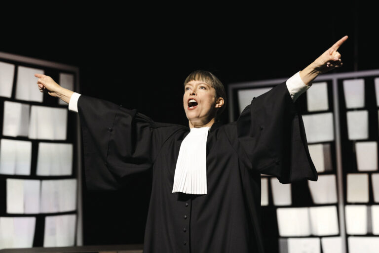 Actrice met rechterstoga aan en beide handen in de lucht met op de achtergrond hangende witte papiertjes in metalen frames