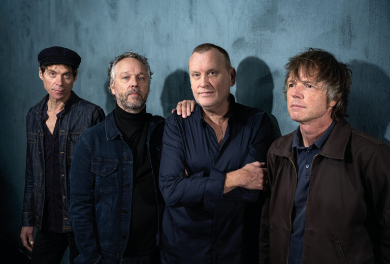 Vier mannen staan voor een blauwe muur in nette donker blauwe kleding en kijken allemaal in de camera