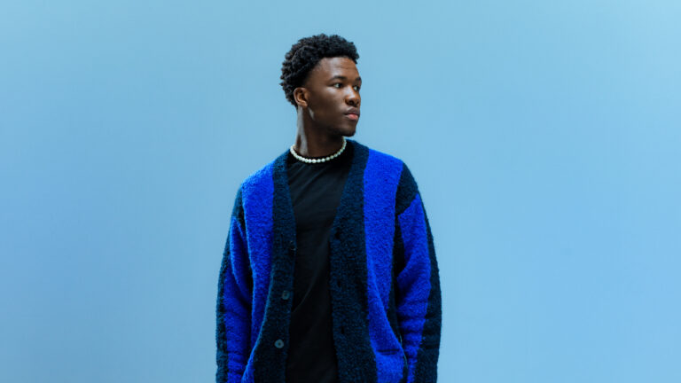 man staat in zwarte outfit met donker blauwe jas voor een blauw achtergrond en kijkt naar de zijkant