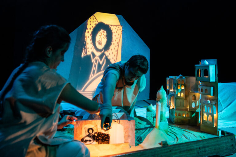 Op het podium wordt er door twee mensen met projecties en gebouwde kleine huizen een verhaal uitgebeeld
