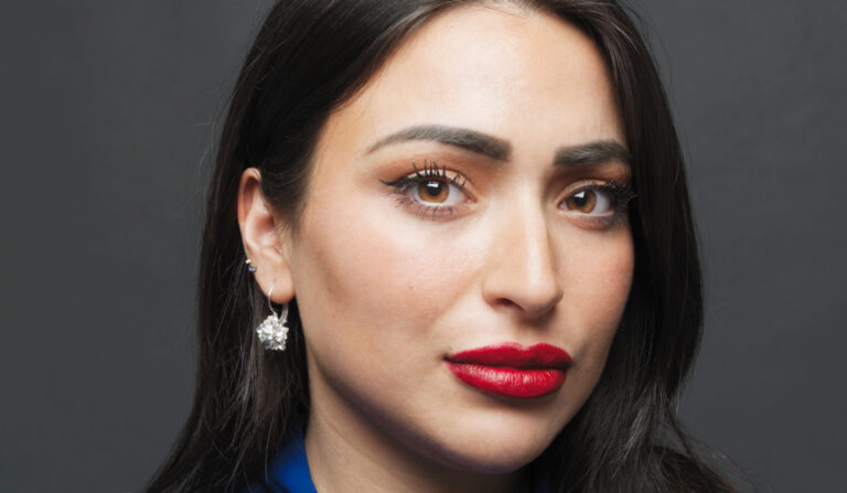 Close-up portret van een vrouw met lang, zwart haar, opvallende make-up en een blauwe kledingstijl.