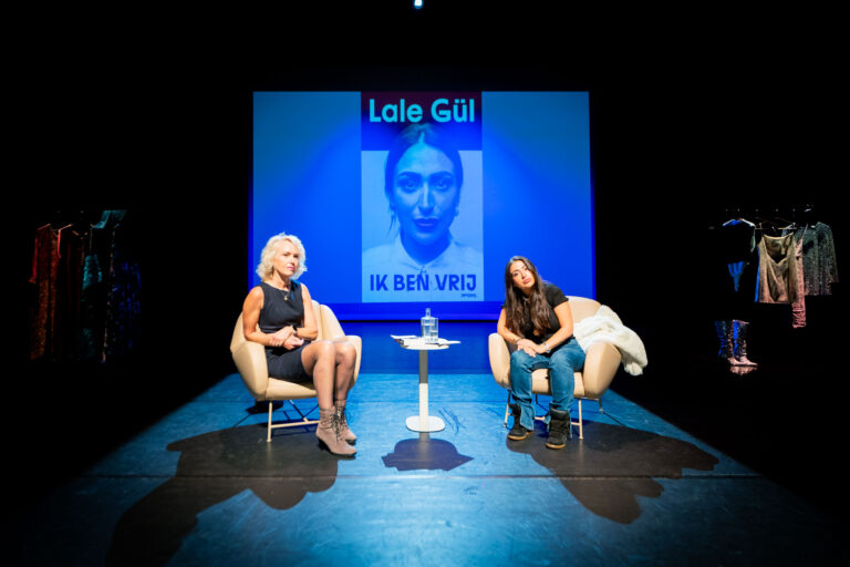 Lale Gül zit met haar interviewer op het podium, voor een geprojecteerd scherm