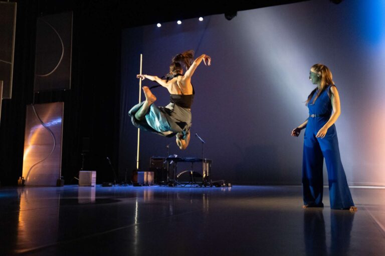 Een danseres springt hoog in de lucht terwijl een andere performer op het podium toekijkt.