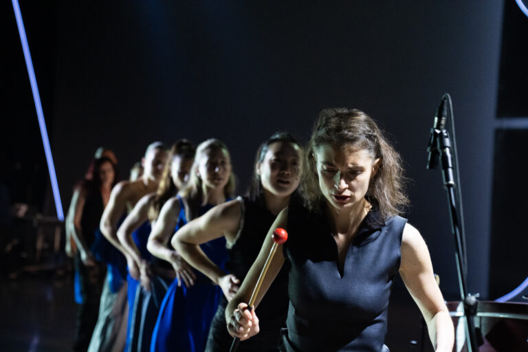 Een rij van vrouwelijke dansers in blauwe en zwarte kleding, geleid door een voorste performer met een mallet in de hand, waarbij zij gefocust en geconcentreerd naar beneden kijkt.