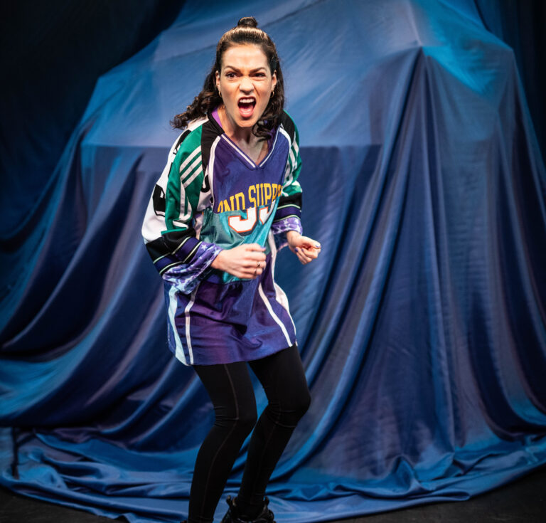 Een actrice in sportkleding staat op een podium en maakt een krachtige, emotionele expressie.