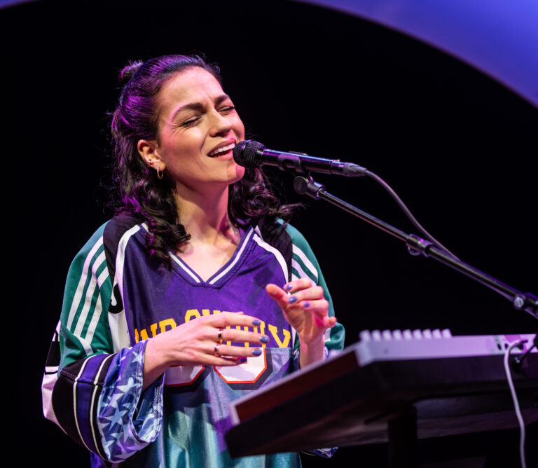 Een vrouwelijke muzikant zingt en speelt een keyboard op een podium, gekleed in een sportieve trui.