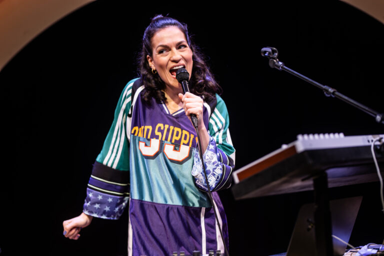 Een vrouw in een kleurrijke sportoutfit staat op een podium en spreekt in een microfoon.