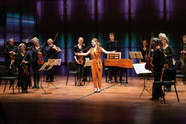 Lucie Horst in een oranje bruine outfit op het podium omringd door staande muzikanten klaar om applaus in ontvangst te nemen