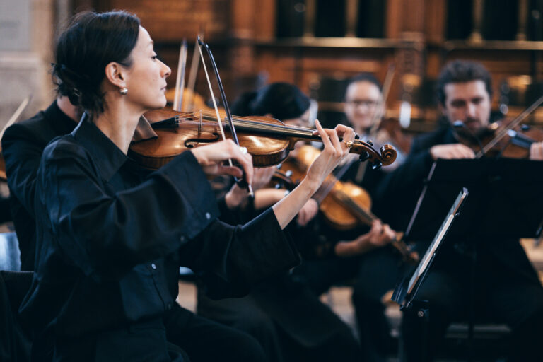 Een vrouw speelt viool als onderdeel van een orkest, omringd door andere muzikanten.