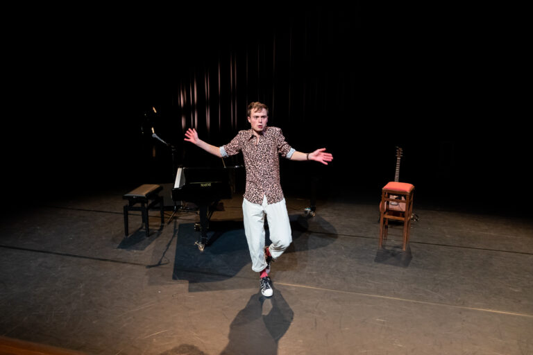 Een performer staat op een podium in een donkere ruimte, met zijn armen wijd open voor een publiek, terwijl een piano en een stoel op de achtergrond staan.