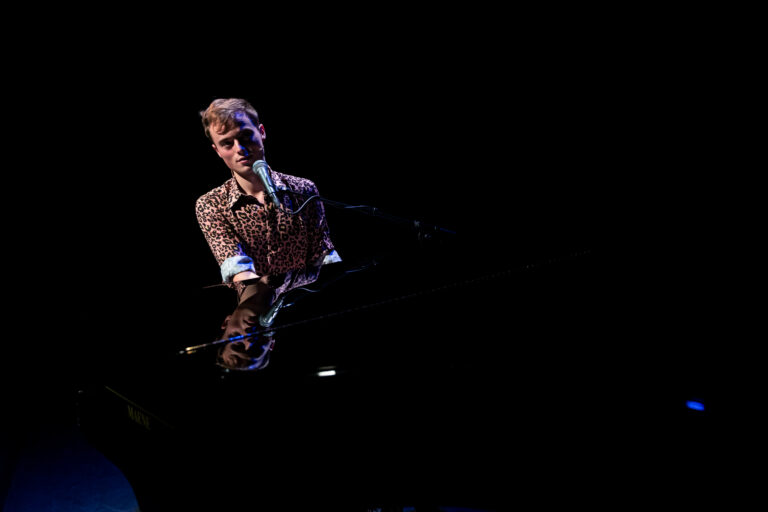 Een muzikant zingt en speelt piano op een goed verlichte podium in een donkere omgeving.
